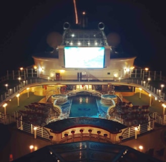 A luxury cruise ship deck featuring a pool surrounded by lounge chairs and warm ambient lighting. A large screen overlooks the area, showing an image against a dark night sky.