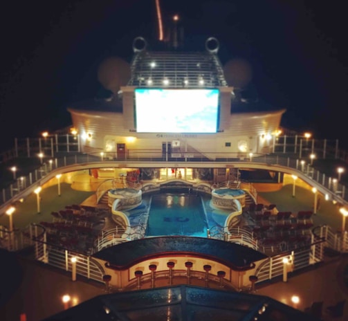 A luxury cruise ship deck featuring a pool surrounded by lounge chairs and warm ambient lighting. A large screen overlooks the area, showing an image against a dark night sky.