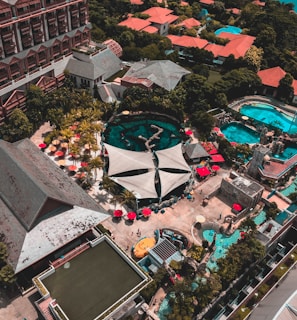 An aerial view of a resort complex with multiple buildings featuring red roofs surrounded by lush greenery. Several swimming pools with turquoise water are visible, alongside poolside areas with red umbrellas and lounge chairs. A large canopy structure is centrally located, and there are walking paths and landscaped gardens throughout the area.