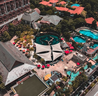 An aerial view of a resort complex with multiple buildings featuring red roofs surrounded by lush greenery. Several swimming pools with turquoise water are visible, alongside poolside areas with red umbrellas and lounge chairs. A large canopy structure is centrally located, and there are walking paths and landscaped gardens throughout the area.