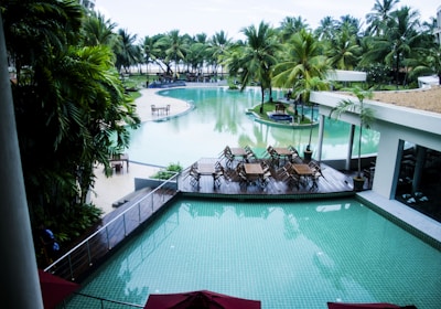 A luxurious resort setting with a large, inviting swimming pool surrounded by palm trees. There is a wooden deck area with dining tables and chairs arranged in a neat fashion. The atmosphere is serene and tropical, with the pool reflecting the greenery of the surrounding trees.