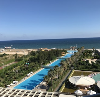 A coastal resort area featuring a long stretch of swimming pools lined with palm trees, adjacent to a clear blue sea. The foreground includes a poolside area with people relaxing under umbrellas, while further towards the horizon, there is a beach with a few parked cars and brightly colored inflatables. The scene is vibrant with lush greenery and well-maintained gardens surrounding the resort facilities.