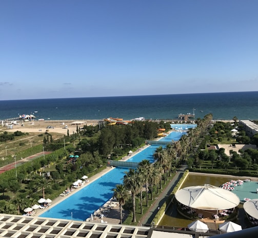 A coastal resort area featuring a long stretch of swimming pools lined with palm trees, adjacent to a clear blue sea. The foreground includes a poolside area with people relaxing under umbrellas, while further towards the horizon, there is a beach with a few parked cars and brightly colored inflatables. The scene is vibrant with lush greenery and well-maintained gardens surrounding the resort facilities.