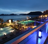 A nighttime scene overlooking a luxurious resort with multiple illuminated swimming pools. The view extends over a body of water with silhouetted islands in the distance. The area is adorned with palm trees and decorative lighting, creating a serene and inviting atmosphere.