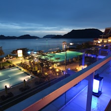 A nighttime scene overlooking a luxurious resort with multiple illuminated swimming pools. The view extends over a body of water with silhouetted islands in the distance. The area is adorned with palm trees and decorative lighting, creating a serene and inviting atmosphere.