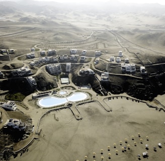 An aerial view of a desert resort complex featuring modern buildings arranged in clusters along winding roads. The center of the development includes two large swimming pools with curved shapes and landscaped areas. The surrounding environment consists of arid, mountainous terrain with sparse vegetation.
