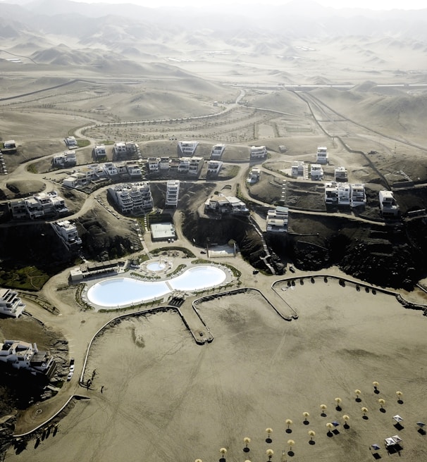 An aerial view of a desert resort complex featuring modern buildings arranged in clusters along winding roads. The center of the development includes two large swimming pools with curved shapes and landscaped areas. The surrounding environment consists of arid, mountainous terrain with sparse vegetation.