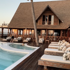 A luxurious wooden resort with a thatched roof, featuring a pool surrounded by lounge chairs and a wooden deck. There are a few people relaxing and walking around the area, and the setting sun casts a warm glow on the building and surroundings.