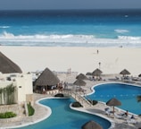 A luxurious beachfront resort features a large pool area with a winding pool bordered by stone pathways and several thatched-roof huts. Sunbeds are placed around the pool, with a few people relaxing under the sun. In the background, a white sandy beach meets a vibrant blue ocean, with waves crashing onto the shore.