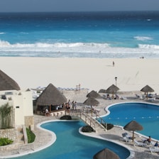 A luxurious beachfront resort features a large pool area with a winding pool bordered by stone pathways and several thatched-roof huts. Sunbeds are placed around the pool, with a few people relaxing under the sun. In the background, a white sandy beach meets a vibrant blue ocean, with waves crashing onto the shore.
