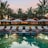 A serene resort setting features a series of lounge chairs lined in a row beside a reflective swimming pool. Large umbrellas provide shade, set against a backdrop of tall palm trees and a warm, glowing structure with expansive windows. The early evening sky casts a soft, calming glow over the entire scene.