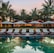 A serene resort setting features a series of lounge chairs lined in a row beside a reflective swimming pool. Large umbrellas provide shade, set against a backdrop of tall palm trees and a warm, glowing structure with expansive windows. The early evening sky casts a soft, calming glow over the entire scene.