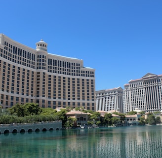 A large luxury hotel and casino building with multiple stories, featuring a light facade with arched windows. The structure has a prominent central tower with a dome-like top. It is surrounded by landscaping with trees and a body of water in the foreground.