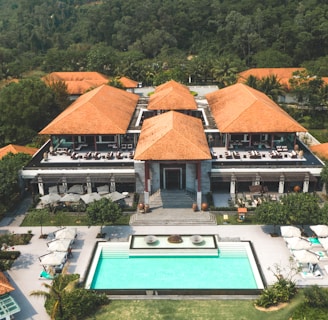 A large resort-style building with several terracotta roofs surrounded by lush green trees and foliage. In front of the building, there is a rectangular swimming pool with sun loungers and umbrellas on a stone patio. The setting is tropical, with palm trees visible around the property, and a forested hillside in the background.