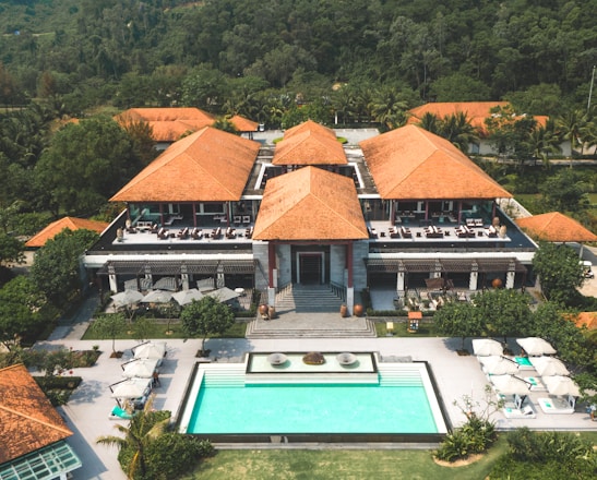 A large resort-style building with several terracotta roofs surrounded by lush green trees and foliage. In front of the building, there is a rectangular swimming pool with sun loungers and umbrellas on a stone patio. The setting is tropical, with palm trees visible around the property, and a forested hillside in the background.