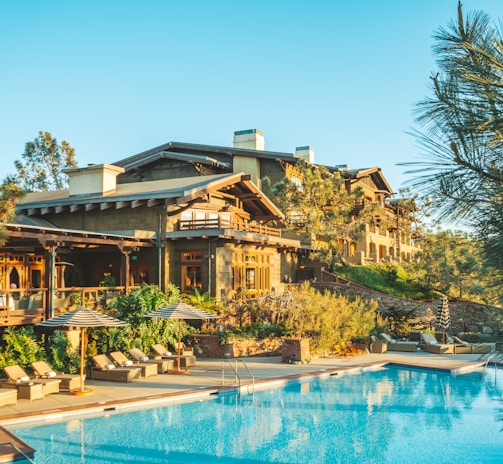 A luxurious resort with wooden architecture surrounded by lush greenery is visible. The main building has a rustic design with multiple levels and balconies. In front of the building, there is a large, inviting swimming pool with lounge chairs and umbrellas arranged neatly beside it. Tall trees and shrubs enhance the natural look of the area, and the sky is clear and blue.