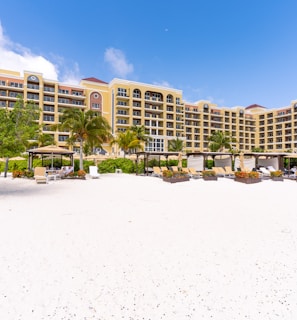 A beachfront resort with a large, elegant building featuring multiple stories and balconies, surrounded by palm trees. The foreground consists of a white sandy beach with scattered lounge chairs and umbrellas. The sky is clear with a few clouds, indicating a sunny day.