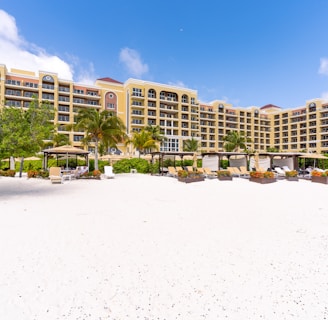 A beachfront resort with a large, elegant building featuring multiple stories and balconies, surrounded by palm trees. The foreground consists of a white sandy beach with scattered lounge chairs and umbrellas. The sky is clear with a few clouds, indicating a sunny day.