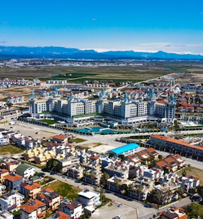 Aerial view of a complex resort situated amid a sprawling urban and suburban area. The resort has expansive buildings with ornate architecture, surrounded by pools and landscaped areas. The surrounding neighborhood consists of residential buildings with a mix of traditional and contemporary styles. Beyond the buildings, the landscape transitions into open fields and distant mountains under a clear blue sky.