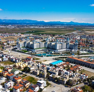 Aerial view of a complex resort situated amid a sprawling urban and suburban area. The resort has expansive buildings with ornate architecture, surrounded by pools and landscaped areas. The surrounding neighborhood consists of residential buildings with a mix of traditional and contemporary styles. Beyond the buildings, the landscape transitions into open fields and distant mountains under a clear blue sky.