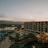 An overview of a resort or hotel complex with a large modern building featuring numerous balconies. In the foreground, there are palm trees and a swimming pool area with lounge chairs and blue umbrellas. The background shows a scenic view of mountains under a sky at sunset, displaying a gradient of pink and orange hues.