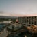 An overview of a resort or hotel complex with a large modern building featuring numerous balconies. In the foreground, there are palm trees and a swimming pool area with lounge chairs and blue umbrellas. The background shows a scenic view of mountains under a sky at sunset, displaying a gradient of pink and orange hues.