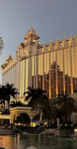 A grand, ornate building with golden illumination stands prominently, reflecting the evening light. Palm trees surround a luxurious pool area, creating an opulent resort atmosphere. The architecture features intricate details and a blend of classic and modern design elements.