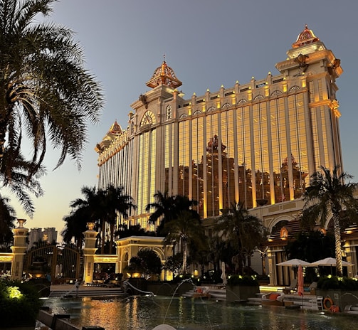 A grand, ornate building with golden illumination stands prominently, reflecting the evening light. Palm trees surround a luxurious pool area, creating an opulent resort atmosphere. The architecture features intricate details and a blend of classic and modern design elements.