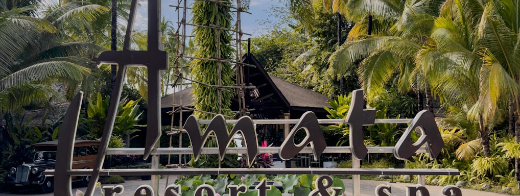 A resort sign is prominently displayed, surrounded by lush tropical plants and palm trees. The sky is blue with scattered clouds, creating a serene atmosphere.
