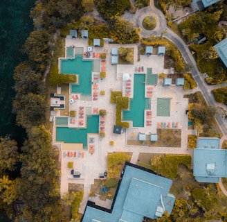 An aerial view of a resort featuring multiple swimming pools with surrounding lounge chairs and umbrellas arranged in a geometric pattern. The area is bordered by lush greenery and pathways leading to various buildings and structures.