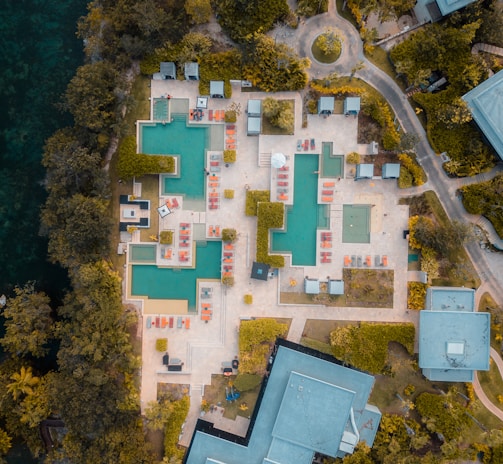 An aerial view of a resort featuring multiple swimming pools with surrounding lounge chairs and umbrellas arranged in a geometric pattern. The area is bordered by lush greenery and pathways leading to various buildings and structures.