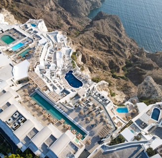 An aerial view of a luxurious coastal resort with multiple swimming pools. The resort features white buildings with terraces and sun loungers, nestled into rocky cliffs overlooking a blue ocean. The landscape is a blend of architectural elegance and natural beauty.