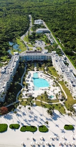 Aerial view of a large resort complex surrounded by lush greenery. The centerpiece is a large swimming pool lined with lounge chairs, nestled within well-maintained gardens. The structures are built in a U-shape, creating an inviting central area. Adjacent to the resort is a pristine white sandy beach with additional sun loungers and shaded areas, indicating a tropical setting.