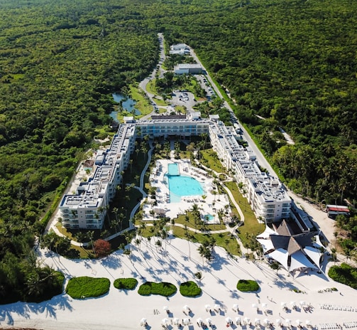 Aerial view of a large resort complex surrounded by lush greenery. The centerpiece is a large swimming pool lined with lounge chairs, nestled within well-maintained gardens. The structures are built in a U-shape, creating an inviting central area. Adjacent to the resort is a pristine white sandy beach with additional sun loungers and shaded areas, indicating a tropical setting.