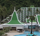 A resort complex featuring a distinctive architectural style with green roofs and white walls, set against a backdrop of lush green hills. The foreground includes palm trees and a large swimming pool, creating a tropical atmosphere.