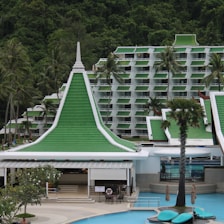 A resort complex featuring a distinctive architectural style with green roofs and white walls, set against a backdrop of lush green hills. The foreground includes palm trees and a large swimming pool, creating a tropical atmosphere.